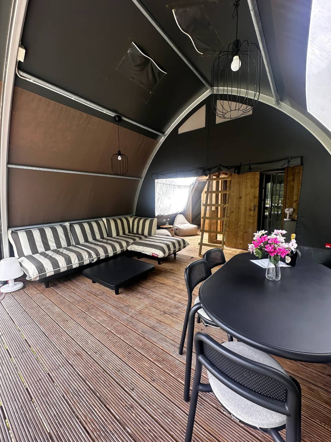 Living area of the Big Oak Lodge XL with a striped lounge sofa, a black dining table with flowers and a wooden ladder