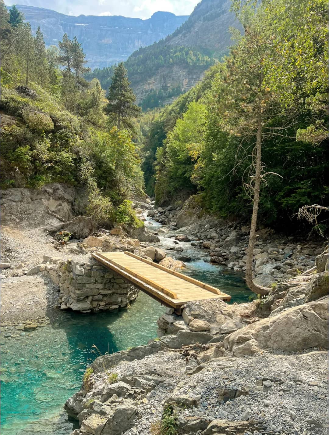 Bright blue mountain river with a wooden platform in a wooded valley surrounded by rocks and conifers, mountains in the background.